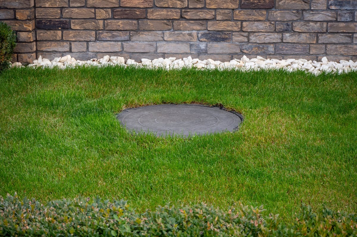 Round utility access cover embedded in green lawn near stone wall. Landscape design, property maintenance, hidden infrastructure. Real photo.