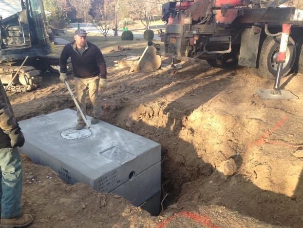 Workers installing a large concrete septic tank into an excavated ground trench at a construction site