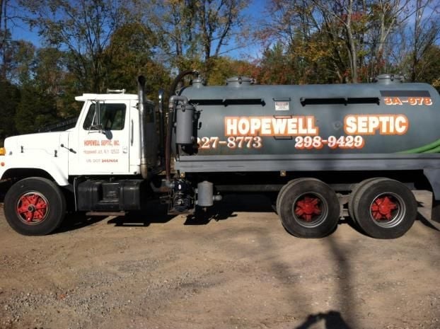 White and dark gray septic truck with red wheels parked on dirt ground with trees in background
