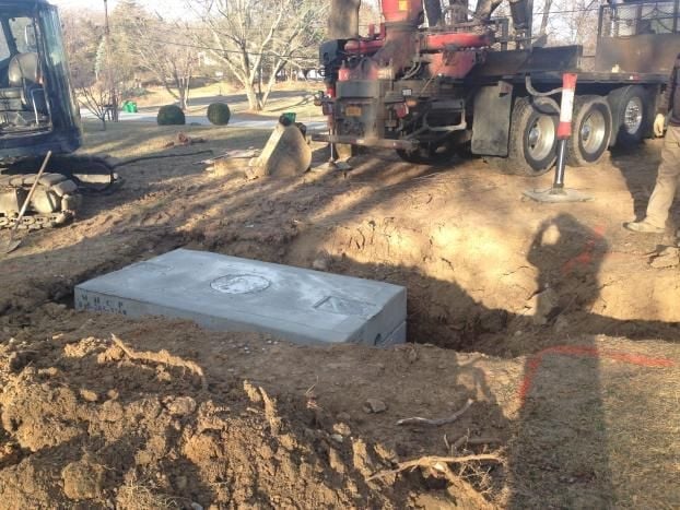Concrete septic tank positioned in excavated ground with construction equipment and trucks in background on residential property