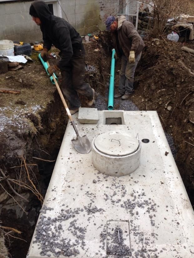 Two workers installing a septic tank system in a dug-out ground trench with pipes and equipment visible