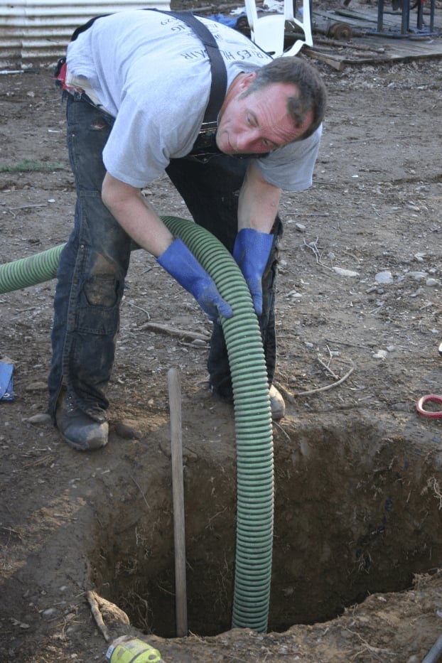 Worker in blue gloves guiding a green corrugated drainage pipe into a hole in the ground at a construction site
