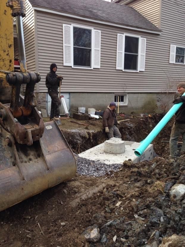 Excavator and workers installing drainage pipe near house foundation during construction project