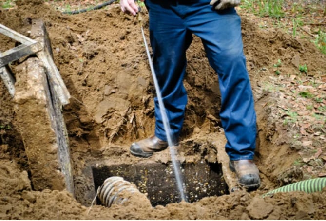 Person in blue jeans digging in soil with a shovel near a metal pipe and drainage system